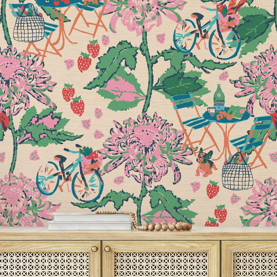 Printed grasscloth wallpaper inspired by the french market, off white base with linear pink florals arranged in vertical stripes.  filled in between are tiny scenes of bicycles with a basket full of market groceries, a bistro set of furniture featuring a bottle of pellegrino and glasses; mixed fruit scattered throughout the print. food fruit bistro chair table